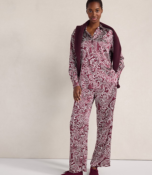 A woman wearing a maroon and white paisley print pajama set with a matching cardigan draped over her shoulders stands against a plain, light-colored wall.