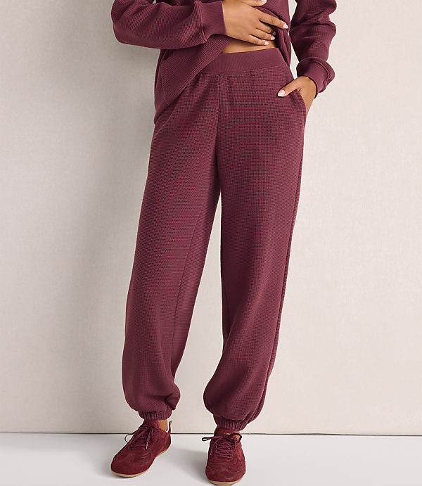 A person wearing a maroon textured lounge set and matching sneakers stands against a plain, light-colored wall.