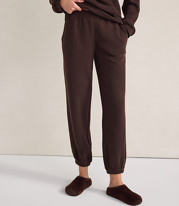 A person wearing brown waffle-knit lounge pants and fuzzy slippers stands against a plain, light-colored wall.
