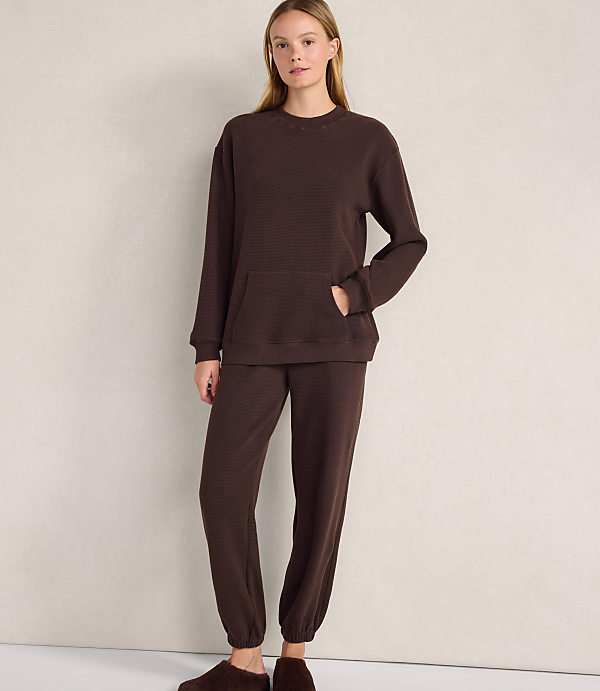 A woman wearing a brown lounge set and slippers stands against a plain, light-colored wall.