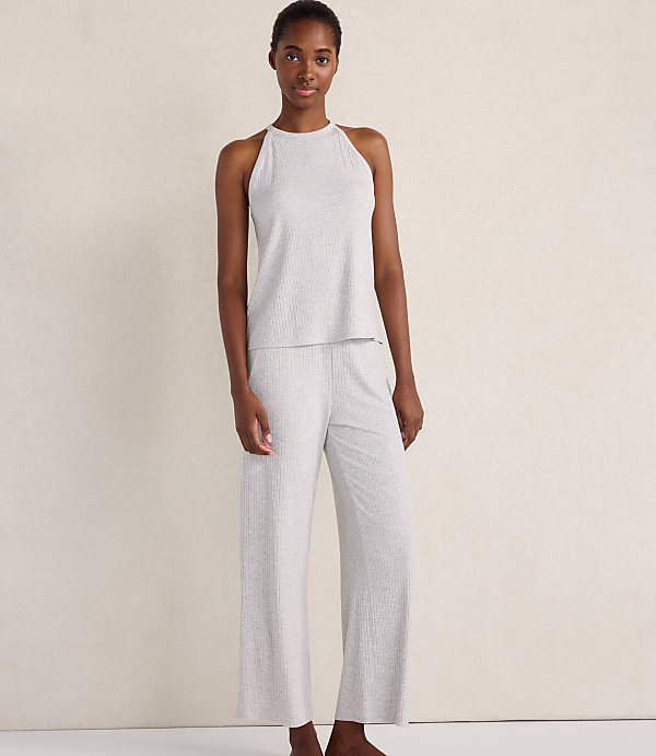 A woman wearing a light grey ribbed sleeveless top and matching wide-leg pants stands against a plain, neutral-toned wall.