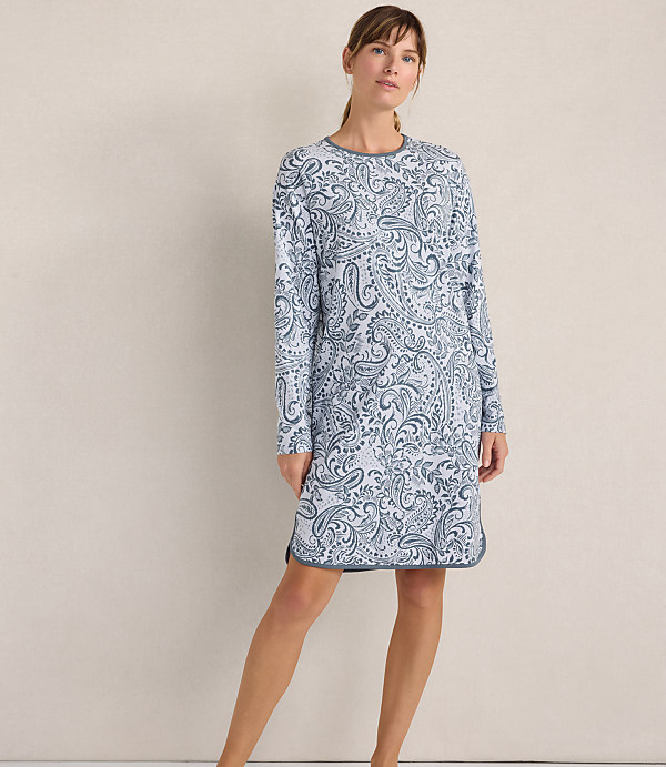 A woman wearing a blue and white paisley print long-sleeved dress stands against a plain, neutral-colored wall.