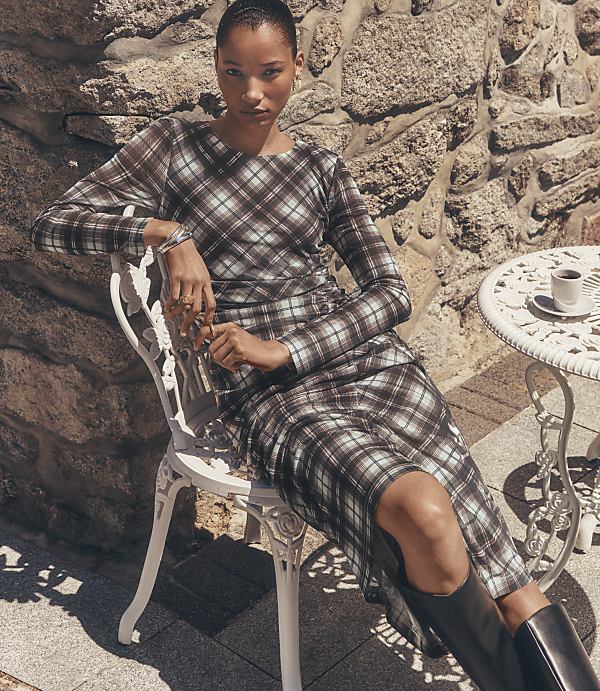 A woman in a plaid dress sits on a white chair in front of a stone wall, with a small table and coffee cup to her right.