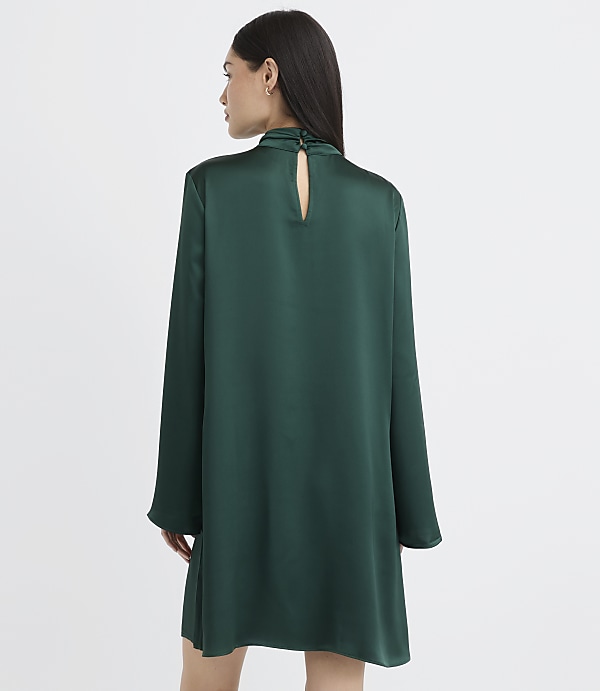 A woman wearing a long-sleeved, emerald green satin dress stands with her back to the camera against a plain white background.