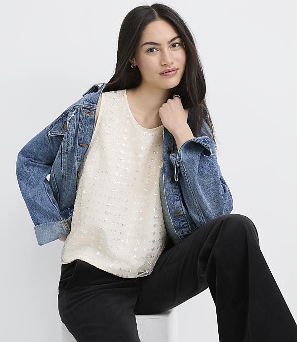 A woman wearing a white sequined top, a blue denim jacket, and black pants sits against a plain white background.