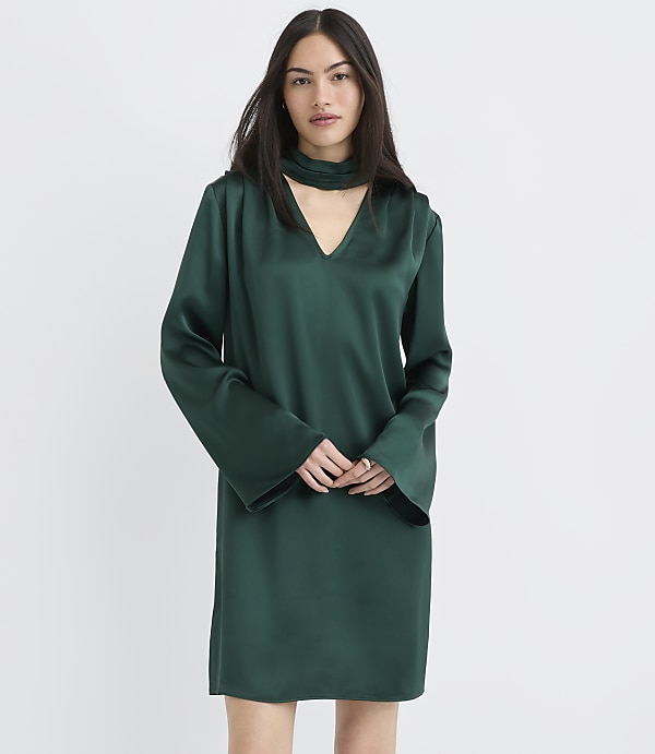 A woman wearing a long-sleeved, dark green satin dress stands against a plain white background.