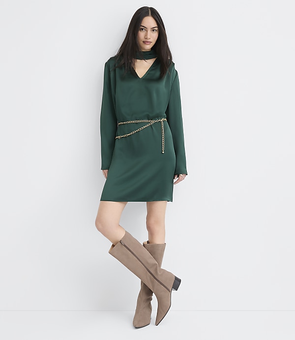 A woman wearing a green satin dress with a gold chain belt and taupe boots stands against a plain white background.