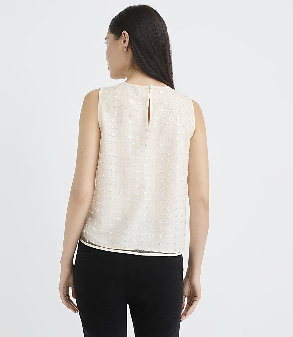 A woman wearing a sleeveless white textured top and black pants stands against a plain white background.