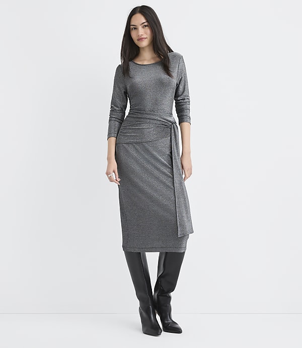 A woman wearing a shimmering silver midi dress and black knee-high boots stands against a plain white background.