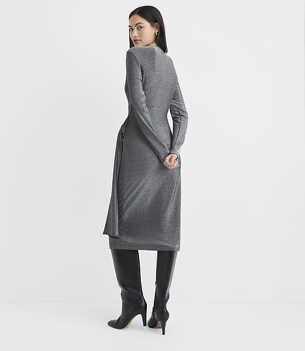 A woman wearing a shimmering silver midi dress and black knee-high boots stands against a plain white background.