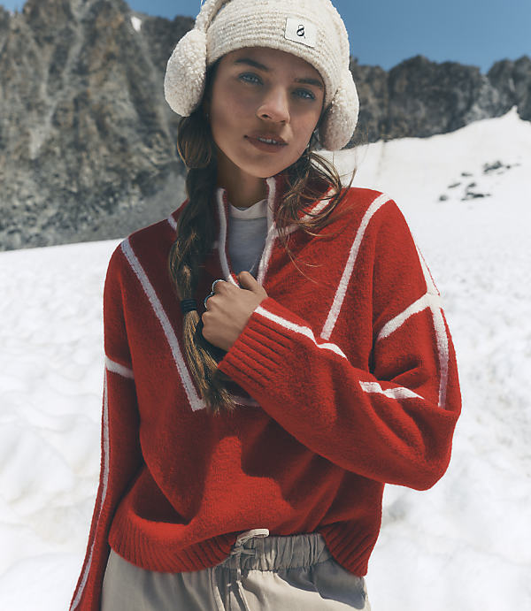 A woman wearing a red patterned sweater and a white beanie stands against a backdrop of snowy slopes and rugged mountains.