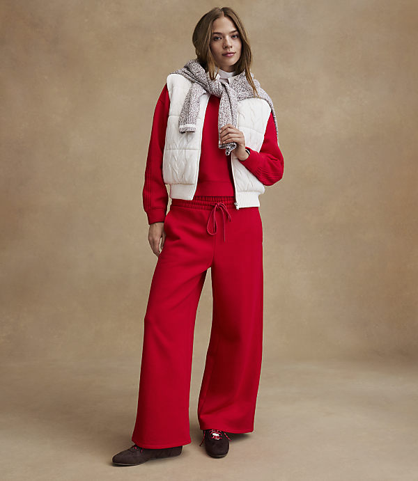 A woman wearing a red outfit with a white vest and grey sweater stands against a neutral, textured tan background.