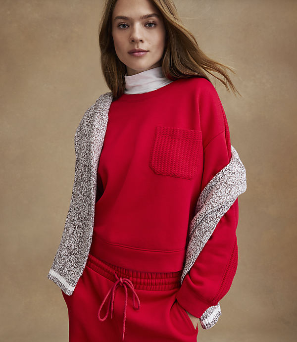 A woman wearing a red sweatshirt and matching pants with a grey cardigan draped over her shoulders stands against a plain tan background.