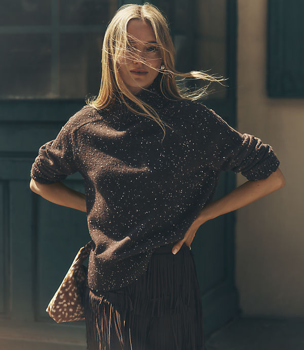 A blonde woman in a dark, speckled sweater and fringed skirt stands with her hands on her hips in front of a dark teal door.