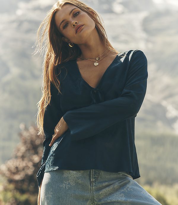 A woman in a dark teal blouse and light blue jeans stands in the foreground against a blurred backdrop of rugged mountains.