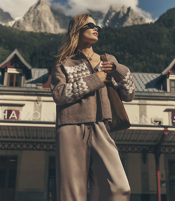 A woman in a patterned brown cardigan and taupe trousers stands in the foreground against a backdrop of a mountain lodge and jagged, snow-capped peaks.
