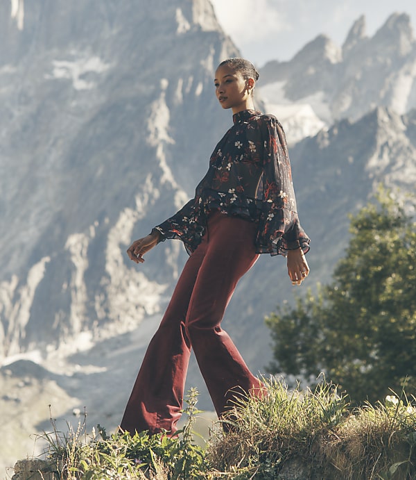 A woman in a floral blouse and red trousers stands on a grassy ledge against a backdrop of towering, jagged mountains.