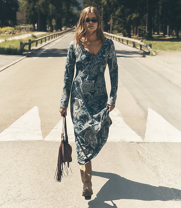 A woman wearing a long, patterned blue dress and sunglasses walks down a sunny road lined with trees and wooden railings.