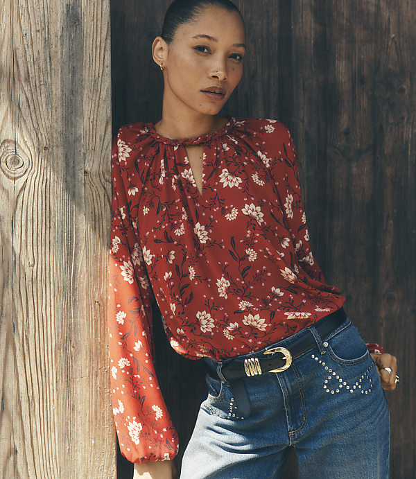 A woman wearing a red floral blouse and blue jeans leans against a rustic wooden wall.