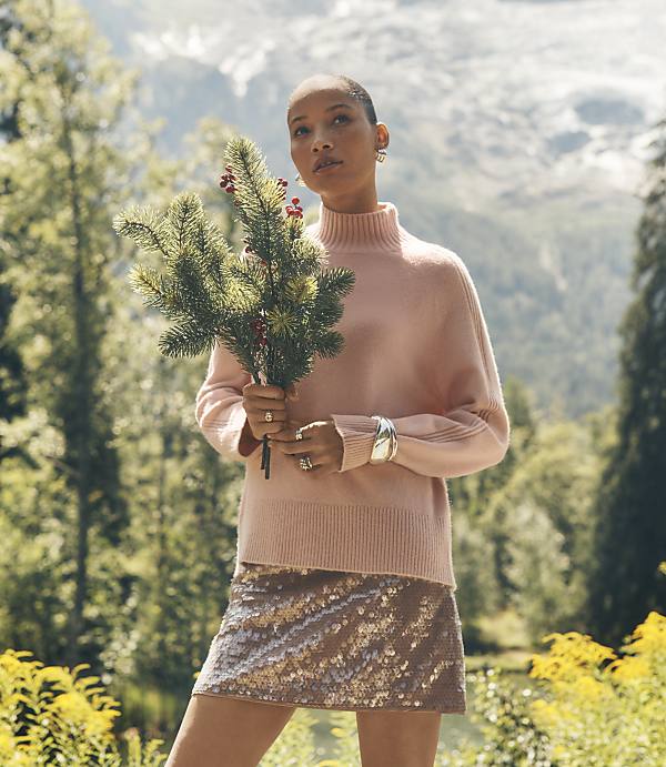 A woman in a pink sweater and sequined skirt holds pine branches against a backdrop of lush green trees and distant mountains.