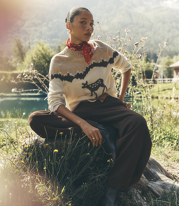 A woman wearing a cow-patterned sweater and brown trousers sits on a mossy rock in a lush landscape with a lake and mountains in the background.