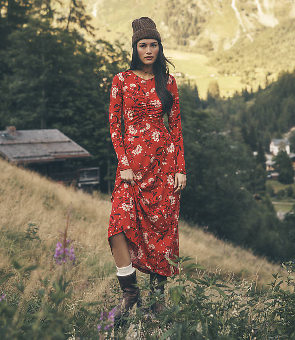 A woman wearing a red floral dress and a brown beanie stands in a grassy field with a rustic cabin and forested mountains in the background.