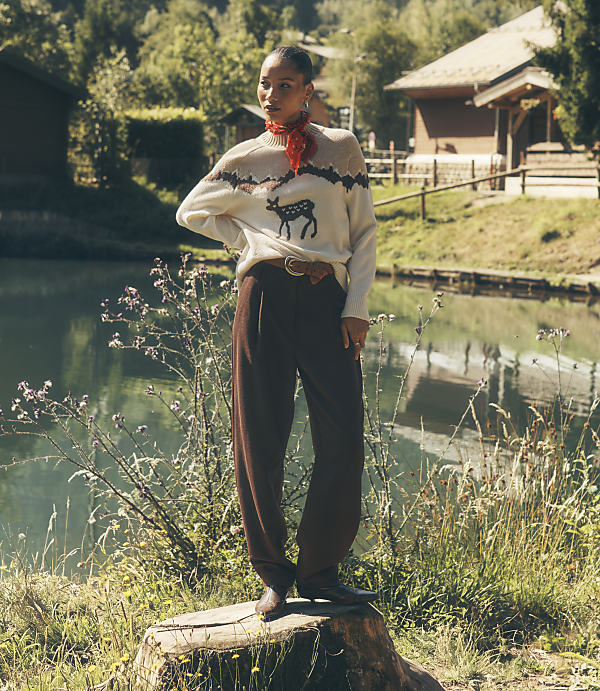 A woman wearing a deer-patterned sweater and brown trousers stands on a tree stump in the foreground, with a calm pond and rustic houses in the background.