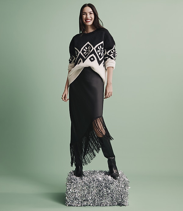 A smiling woman wearing a patterned sweater, black skirt, and boots stands on a silver foil pedestal against a solid light green background.