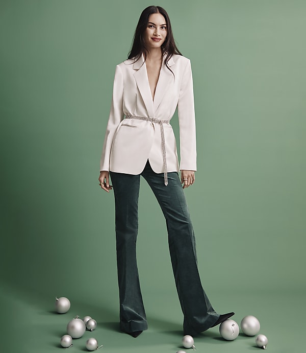 A woman wearing a white blazer and dark green trousers stands among silver ornaments against a solid green background.