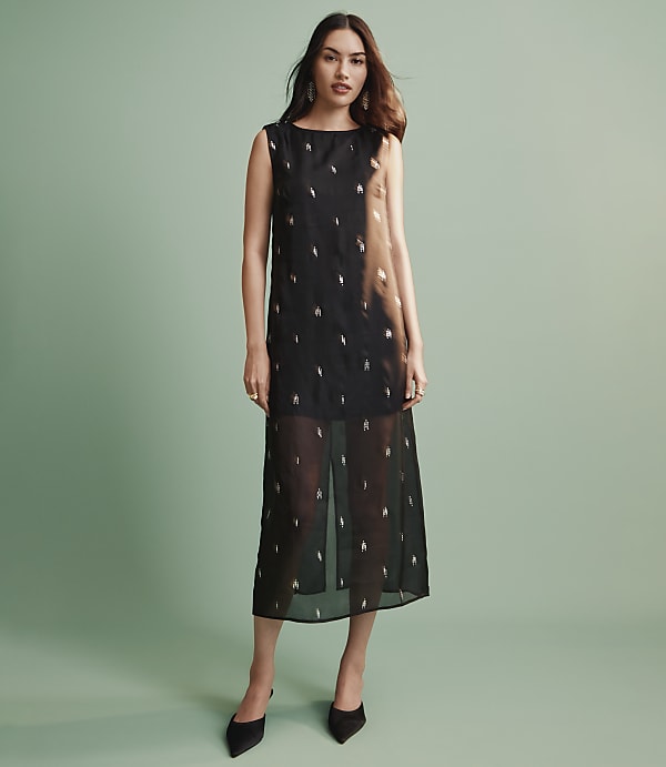 A woman wearing a sleeveless black midi dress with metallic accents and black pointed heels stands against a solid light green background.