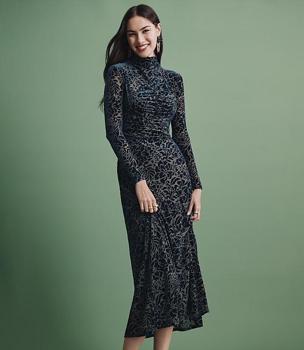 A smiling woman wearing a long-sleeved, dark teal patterned dress stands against a solid sage green background.