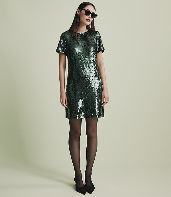 A woman wearing a green sequined dress, black tights, and sunglasses stands against a plain light green background.