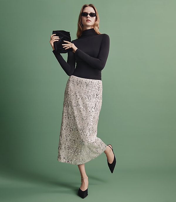 A woman wearing a black turtleneck, a shimmering silver sequined skirt, and sunglasses stands against a solid green background.