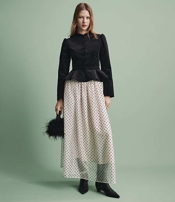 A woman wearing a black velvet peplum top and a sheer polka-dot skirt stands against a solid light green background.
