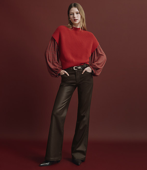 A woman wearing a red sweater and brown flared trousers stands against a solid red background.