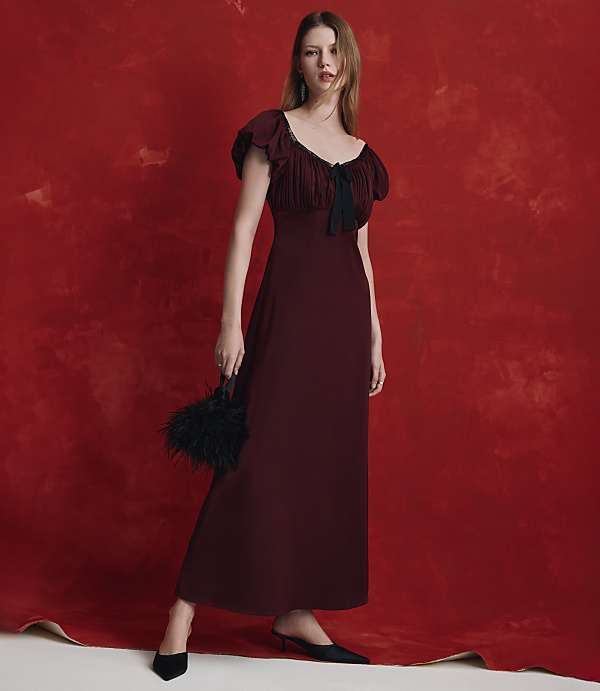 A woman in a long burgundy dress and black heels stands against a textured red backdrop.