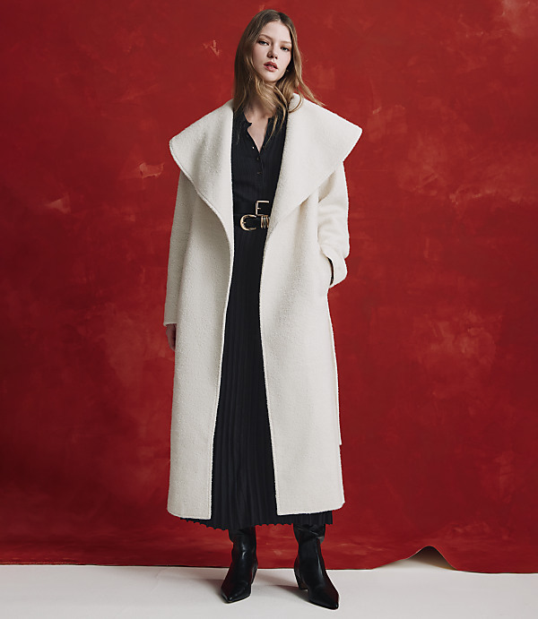 A woman wearing a long white coat over a black pleated dress and black boots stands against a textured red background.