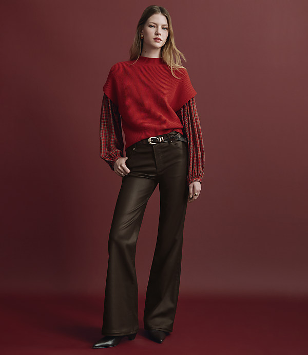 A woman wearing a red layered top and brown flared trousers stands against a solid red background.