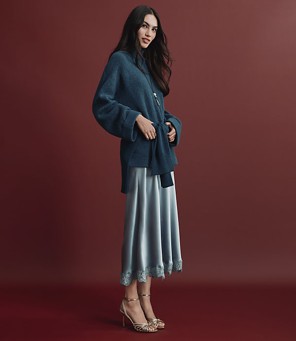 A woman wearing a teal knit sweater and a shimmering light blue satin skirt stands against a solid deep red background.