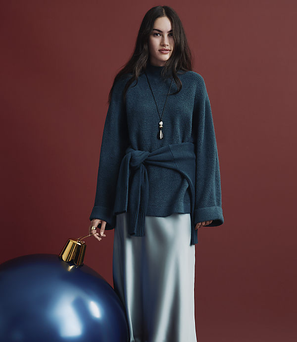 A woman wearing a teal knit sweater and a satin skirt stands next to a giant blue ornament against a solid red background.