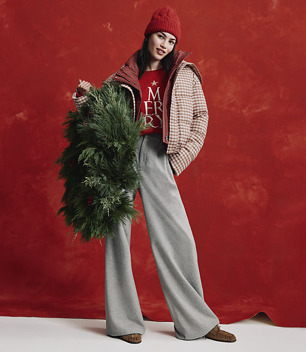 A woman wearing a red beanie, a checkered jacket, and wide-leg grey trousers holds a large evergreen wreath against a textured red background.