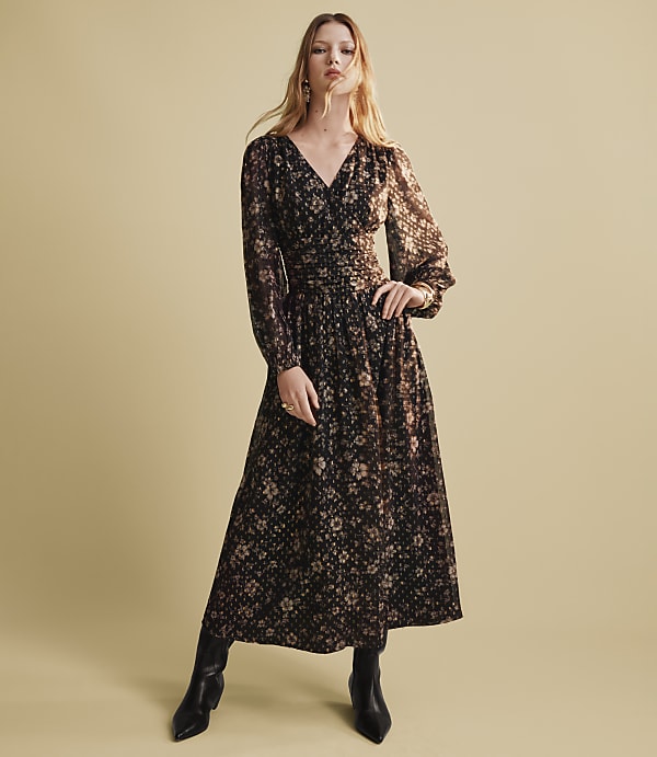 A woman wearing a long-sleeved floral maxi dress and black boots stands against a plain, light beige background.