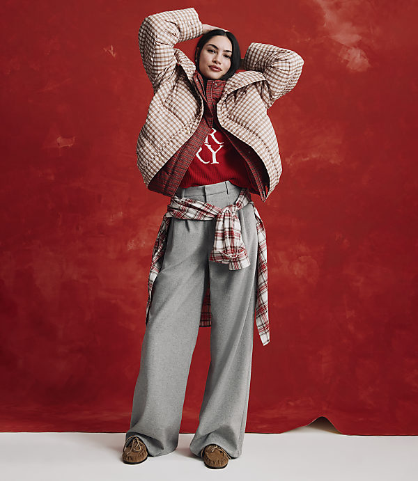 A woman wearing a checkered jacket, red sweater, and wide-leg grey trousers stands against a textured red background.