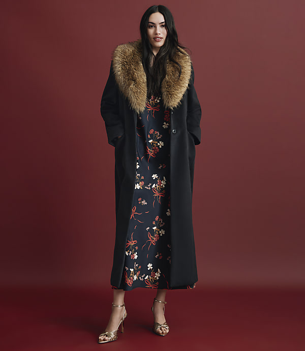 A woman wearing a long black coat with a fur collar over a floral dress and gold heels stands against a solid red background.