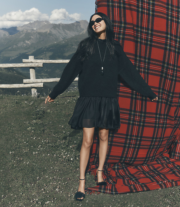 A woman in a black outfit stands before a large red plaid backdrop on a grassy hill with mountains in the distance.