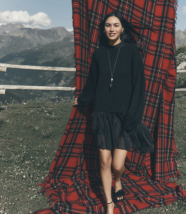 A woman in a black sweater and skirt stands before a large red plaid blanket with a mountainous landscape in the background.