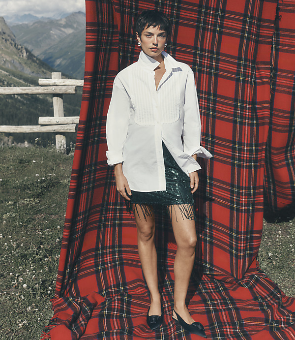 A woman wearing a white shirt and a green sequined skirt stands before a large red plaid backdrop with a mountainous landscape in the distance.