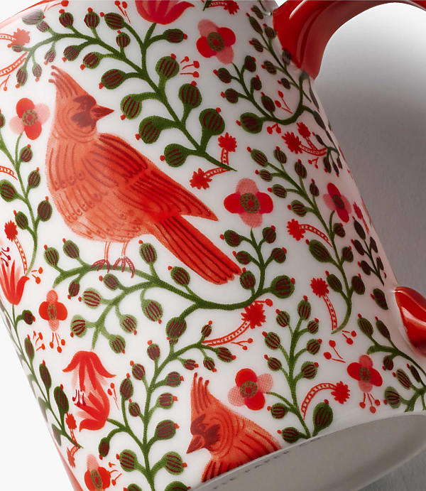 A red cardinal and floral patterns decorate the foreground of a white ceramic mug against a plain white background.