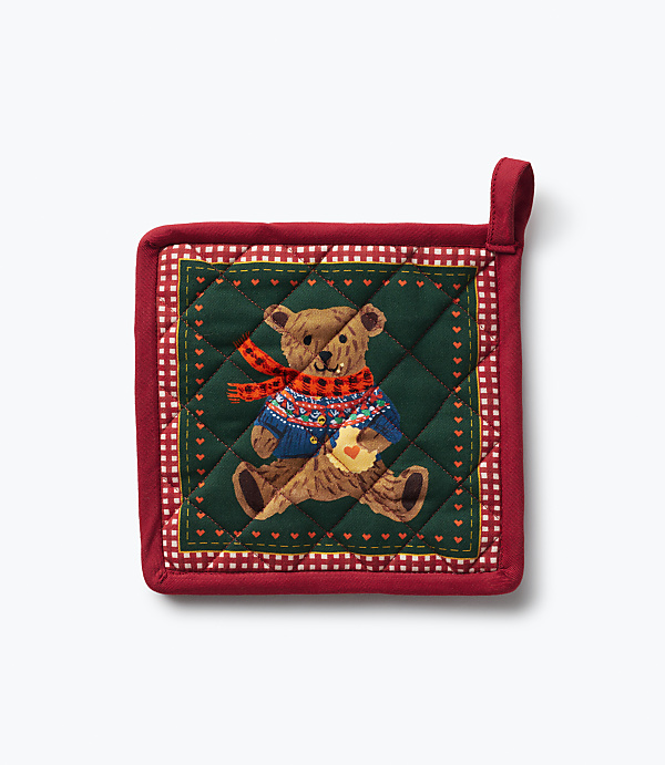 A red quilted pot holder featuring a teddy bear in a sweater and scarf sits against a plain white background.