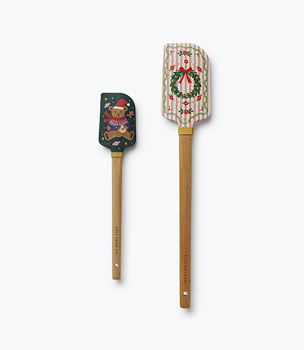 Two decorative spatulas, one featuring a teddy bear and the other a holiday wreath, are positioned vertically against a plain white background.